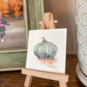 Puede incluir: Una acuarela de una calabaza turquesa sobre un pequeño caballete de madera. La calabaza tiene un tallo curvo y está sobre un fondo claro. El caballete está sobre una superficie de madera, con una foto enmarcada y una maceta de cerámica blanca en el fondo.