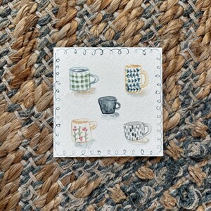 May include: Watercolor painting of five mugs with various patterns and colors, including green plaid, blue and yellow designs, a solid black mug, and floral and abstract patterns. The artwork is on a white square with a decorative border, set against a textured, woven background.