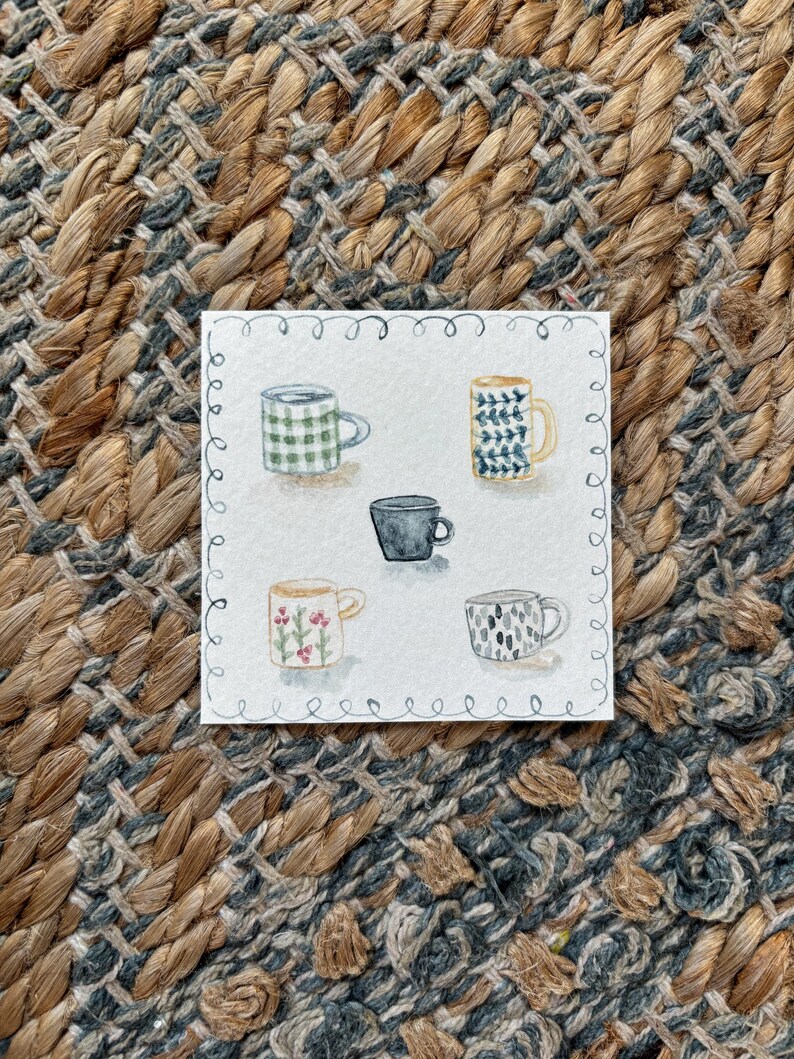 May include: Watercolor painting of five mugs with various patterns and colors, including green plaid, blue and yellow designs, a solid black mug, and floral and abstract patterns. The artwork is on a white square with a decorative border, set against a textured, woven background.