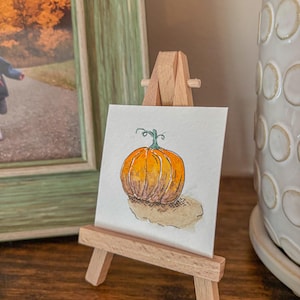 Puede incluir: Una acuarela de una calabaza naranja con un tallo verde, expuesta en un pequeño caballete de madera. La obra de arte se encuentra sobre un fondo neutro, lo que sugiere un tema de otoño o cosecha. La pintura está detallada con sombras y luces.