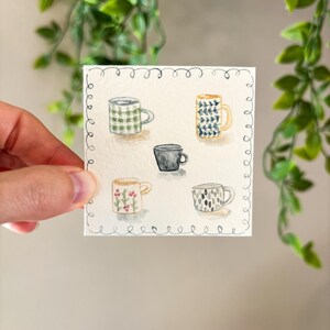 May include: Watercolor painting of five mugs with various designs. The mugs include a green plaid pattern, a blue and white floral pattern, a solid black mug, a mug with pink flowers, and a mug with a black and white pattern. The artwork is on a square piece of paper.
