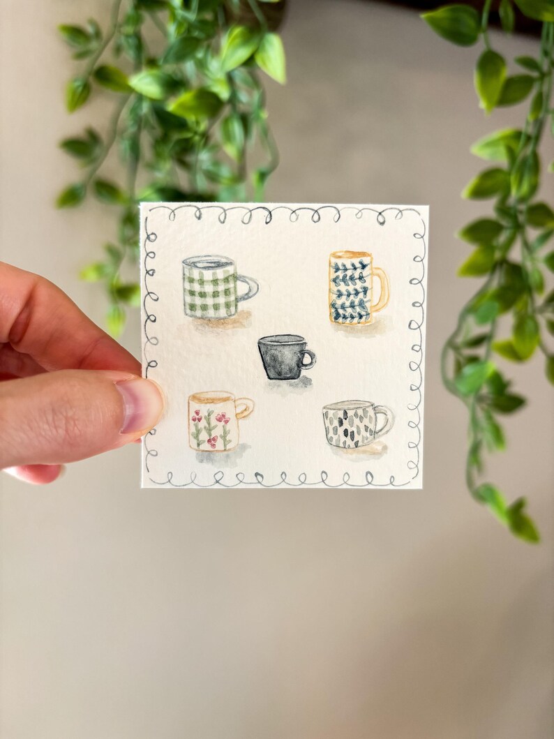 May include: Watercolor painting of five mugs with various designs. The mugs include a green plaid pattern, a blue and white floral pattern, a solid black mug, a mug with pink flowers, and a mug with a black and white pattern. The artwork is on a square piece of paper.