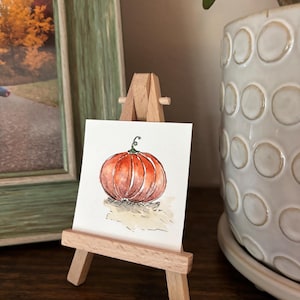 Puede incluir: Una acuarela de una calabaza naranja con un tallo verde, exhibida en un pequeño caballete de madera. La obra de arte se presenta sobre un fondo neutro, sugiriendo un tema de otoño o cosecha. La calabaza es el punto focal.