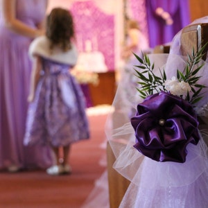 May include: A purple and white tulle chair decoration with a large purple satin flower and white pearl accents.