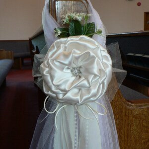 May include: A white satin flower with a pearl embellishment, tied with a white tulle ribbon and white satin ribbon, for a wedding ceremony decoration.