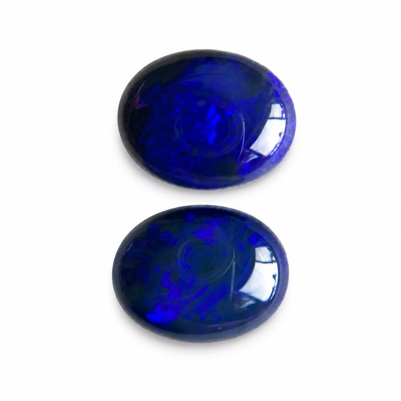 May include: Two oval-shaped, polished gemstones with a deep blue color and internal patterns. The gems are set against a white background, showcasing their vibrant hues and reflective surfaces. The stones appear to be approximately 1 inch in diameter.