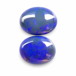 May include: Two oval-shaped opals with a polished, glossy finish. The opals display a vibrant blue color with internal flashes of purple and hints of other colors. The gemstones are set against a white background.