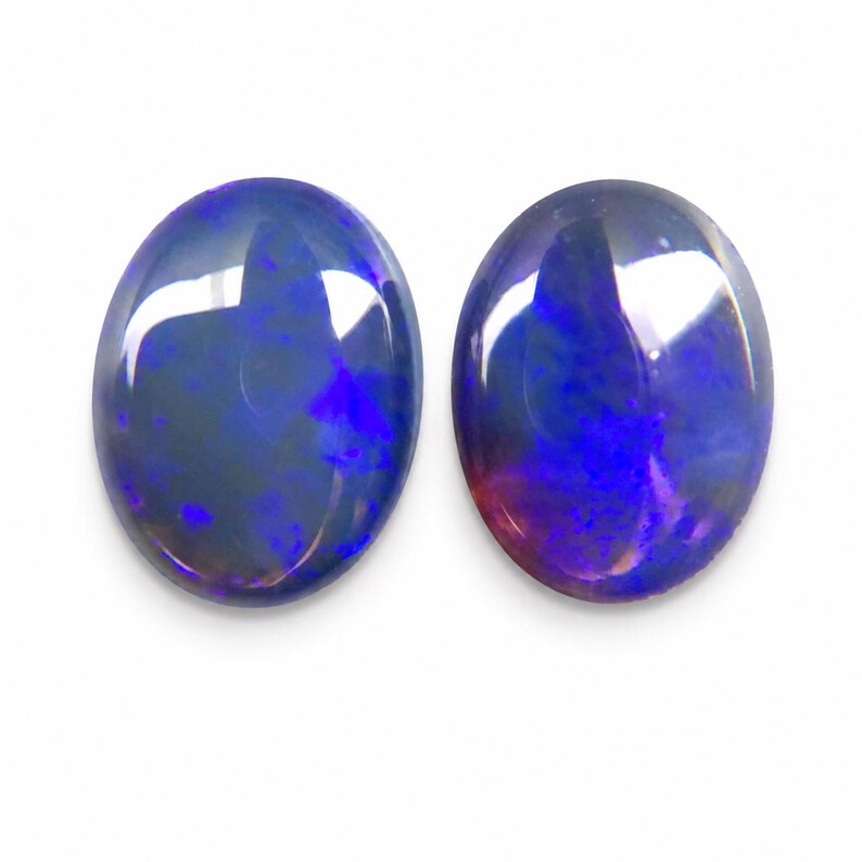 May include: Two oval-shaped opals with a dark blue base color and vibrant flashes of blue and purple. The gemstones are polished and have a glossy appearance, set against a white background.