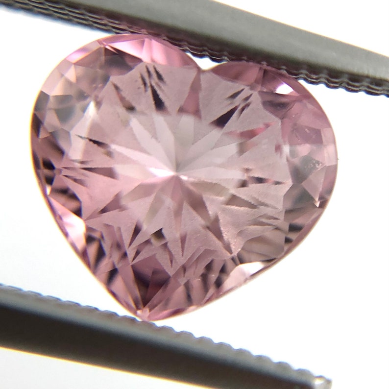 May include: A heart-shaped, faceted gemstone with a pink hue. The gem is held in tweezers and has a star-like pattern within the stone.