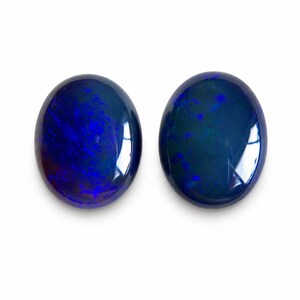 May include: Two oval-shaped, polished gemstones with a deep blue color and internal patterns. The gems have a glossy surface, reflecting light. The stones are set against a white background.