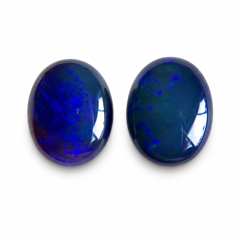 May include: Two oval-shaped, polished gemstones with a deep blue color and internal patterns. The gems have a glossy surface, reflecting light. The stones are set against a white background.