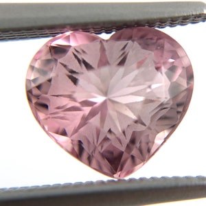 May include: A pink heart-shaped gemstone with a faceted cut. The gemstone is held in tweezers and has a star-like pattern.