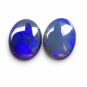 May include: Two oval-shaped opals with a dark blue base color and vibrant blue and purple flashes. The gemstones are polished and reflect light, set against a white background. These opals are suitable for jewelry making.
