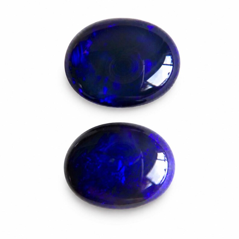 May include: Two oval-shaped, polished gemstones with a deep blue color and internal patterns. The gems have a glossy surface, reflecting light. The background is plain white, highlighting the gemstones' color and shape. These are likely opals.