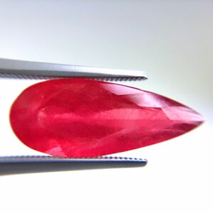 Puede incluir: Una gema facetada en forma de lágrima, de color rojo vibrante. La gema está sujeta por unas pinzas plateadas sobre un fondo blanco brillante. La superficie de la gema refleja la luz, mostrando sus facetas pulidas. Adecuada para la creación de joyas. Mide unos 2.5 cm.