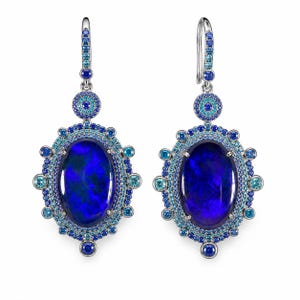May include: A pair of ornate earrings featuring large, oval-shaped, dark blue opals. The opals are surrounded by a halo of smaller blue and turquoise gemstones, with additional accents and a silver hook.