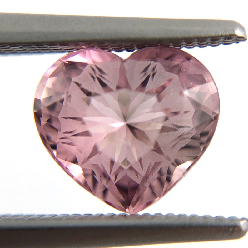 May include: A heart-shaped pink gemstone with a faceted cut. The gemstone is held by tweezers.