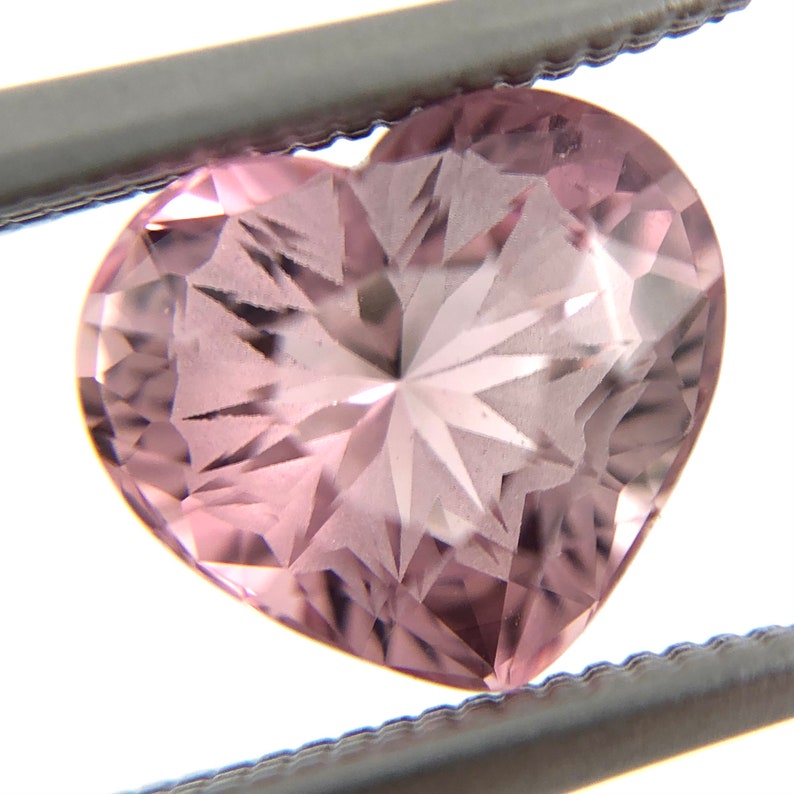 May include: A pink heart-shaped gemstone with a star-shaped pattern inside. The gemstone is held by tweezers.