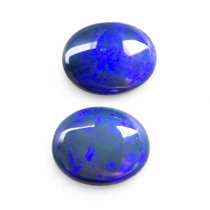 May include: Two oval-shaped opals with a dark blue base color and flashes of vibrant blue and purple. The gemstones are polished and reflect light, set against a white background. These opals are suitable for jewelry making.