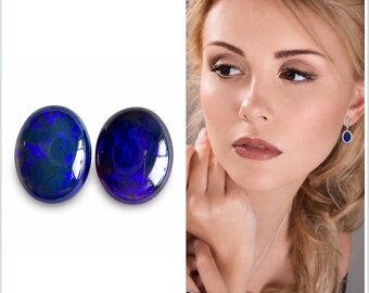 May include: Two oval-shaped opals with a dark blue and green iridescence. The opals are displayed on a white background. The image also shows a woman wearing matching jewelry: earrings and a ring with similar blue gemstones.