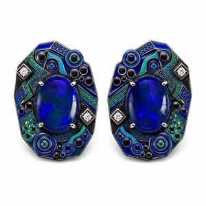 May include: A pair of ornate earrings featuring large, oval, blue gemstones. The earrings have a geometric design with blue, green, black, and white accents. Small diamonds and circular details add to the intricate design. The earrings are set against a white background.