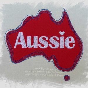 May include: Red felt outline of the continent of Australia with the word "Aussie" embroidered in white with a small white heart above the word. The outline is stitched with blue thread.