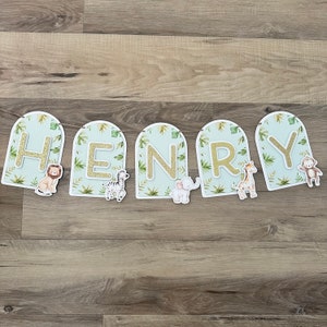 May include: A green and gold banner with the name "HENRY" spelled out in gold letters. Each letter is decorated with a different jungle animal, including a lion, zebra, elephant, giraffe, and monkey.