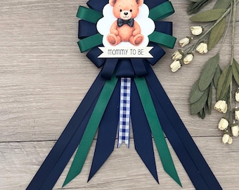 May include: A navy blue, green, and white baby shower rosette with a teddy bear illustration. The rosette has a central image of a teddy bear wearing a bow tie, with the text "mommy to be" on a banner.