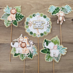 May include: Set of six jungle animal themed cake toppers. The toppers feature a lion, monkey, zebra, giraffe, elephant, and a round topper with the text "A Wild One is on the Way!"