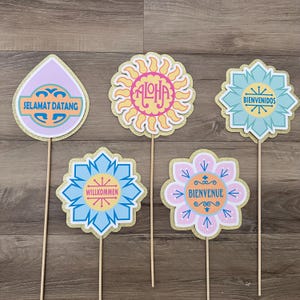 May include: A collection of colorful photo booth props on wooden sticks. The props feature various shapes, including flowers and a teardrop, with phrases like "Aloha" and "Welcome" in different languages. Gold glitter borders add a decorative touch.
