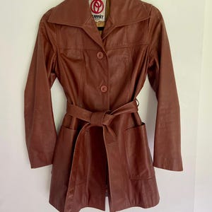 May include: A brown leather jacket featuring a wide collar, long sleeves, and a matching tie belt at the waist. The jacket has two front pockets and two buttons down the centre. A label inside the neck displays the text ANNET.