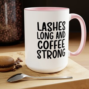 May include: White ceramic coffee mug with a pink rim and handle. The mug has black text that reads "Lashes Long and Coffee Strong".