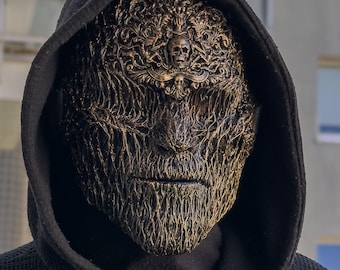 Könnte beinhalten: Eine Vollmaske mit einer texturierten, verwitterten Bronzeoberfläche, die mit komplizierten Filigranmustern und einem zentralen Totenkopf-Emblem versehen ist. Die Maske wird unter einem schwarzen Kapuzenpullover aus strukturiertem Stoff getragen.