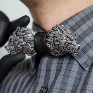 Peut inclure: Un nœud papillon argenté orné de deux sculptures de têtes de loup, chacune avec des détails de fourrure et des expressions menaçantes. Le nœud papillon a une bande en cuir noir et est porté avec une chemise à carreaux grise.