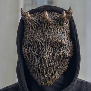 Op de afbeelding: Een volledig gezichtsmasker met een complexe, wortelachtige of webachtige textuur in een donkerbrons en zwarte afwerking. Het masker heeft drie prominente, hoornachtige uitsteeksels aan de bovenkant en wordt gedragen met een zwarte capuchon.