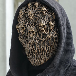 Op de afbeelding: Een volledig gezichtsmasker met een complex netwerk van verhoogde, bronskleurige, wortelachtige structuren verweven met meerdere kleine, gedetailleerde menselijke schedels. Het masker wordt gedragen onder een zwarte capuchon.