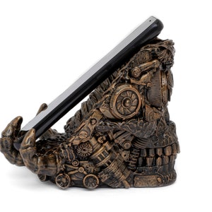 May include: A bronze-colored, steampunk-style phone stand shaped like a hand holding a smartphone. The stand is made of intricate gears and metal parts.