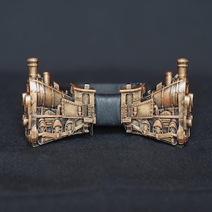 Könnte beinhalten: Eine goldfarbene Fliege in Form einer Lokomotive. Die Fliege ist aus Metall und hat einen schwarzen Lederriemen in der Mitte.