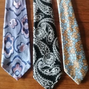 Pode incluir: Três gravatas com padrões diferentes. A primeira gravata é azul com desenhos florais rosa e branco. A segunda gravata é preta com um padrão paisley branco. A terceira gravata é azul claro com desenhos florais castanhos e brancos.