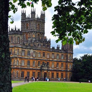 Könnte beinhalten: Ein großes, steingebautes Gebäude mit einem hohen Turm und vielen Fenstern. Das Gebäude ist von einem grünen Rasen und Bäumen umgeben. Vor dem Gebäude gehen Menschen spazieren.