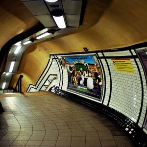 Op de afbeelding: Een gebogen, betegelde gang in een metrostation met een filmposter die "Shaun of the Dead" adverteert en andere advertenties op de muren.