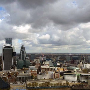 May include: A panoramic view of London's skyline with several skyscrapers, including the Shard, under a cloudy sky.