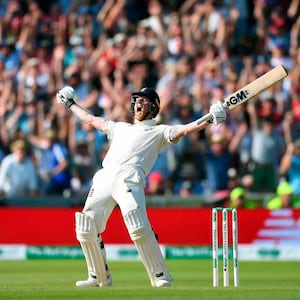 Puede incluir: Un jugador de cricket con un uniforme blanco levanta su bate en el aire después de golpear una bola. Lleva un casco y equipo de protección. El jugador está de pie en un campo verde con una barrera roja y blanca en el fondo. Se puede ver una multitud de personas en el fondo animando.
