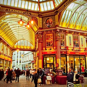 Puede incluir: Imagen del interior del mercado de Leadenhall en Londres. Muestra una escena bulliciosa con gente caminando por los arcos ornamentados. La arquitectura presenta colores rojo, dorado y crema, con techos de cristal y faroles colgantes.