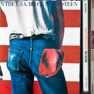Puede incluir: Portada del álbum "Born in the U.S.A." de Bruce Springsteen, que muestra la espalda de un hombre con vaqueros y camisa blanca, con una gorra roja en el bolsillo trasero. El fondo muestra la bandera estadounidense. Se ven otros álbumes de Springsteen.