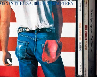 May include: Album cover for Bruce Springsteen's "Born in the U.S.A.", showing a man in blue jeans and a white shirt, with a red cap in his back pocket. The background features the American flag. Other Springsteen album CDs are visible.