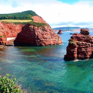 May include: A panoramic view of a rocky coastline with red cliffs and a turquoise ocean. The cliffs are jagged and eroded, creating a dramatic landscape. The water is clear and calm, reflecting the blue sky.