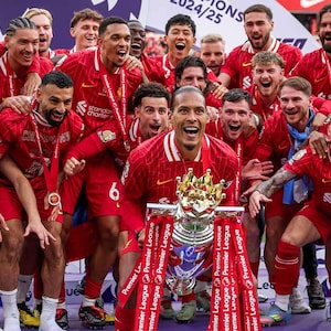 Op de afbeelding: Een groep voetballers in rode shirts viert met een trofee. De trofee is zilver en goud, met rode linten met de tekst "Premier League". De spelers dragen medailles en lachen.