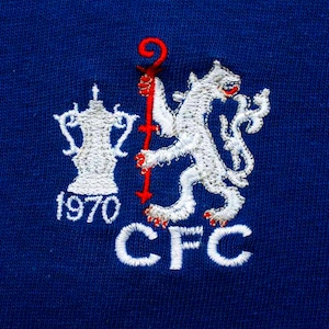 Puede incluir: Un león blanco bordado sosteniendo un bastón rojo con una copa blanca y el texto "1970 CFC" sobre un fondo azul.