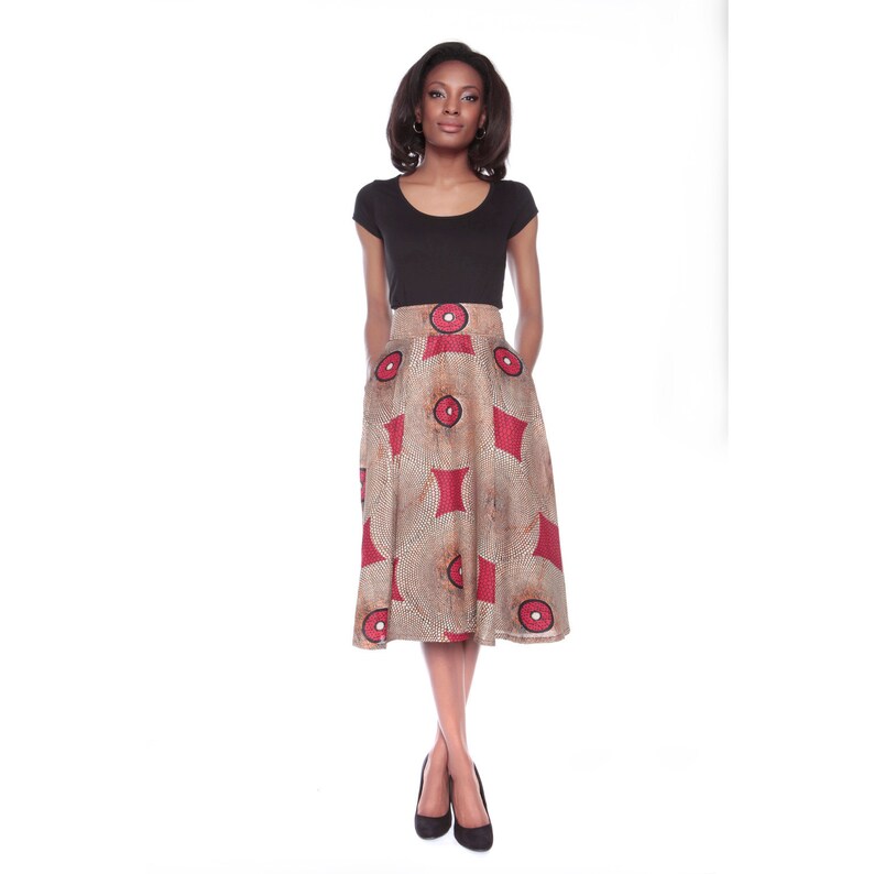 May include: A woman wearing a black short-sleeved shirt and a brown and red patterned skirt with a high waist. She is standing with her hands in her pockets and wearing black heels.